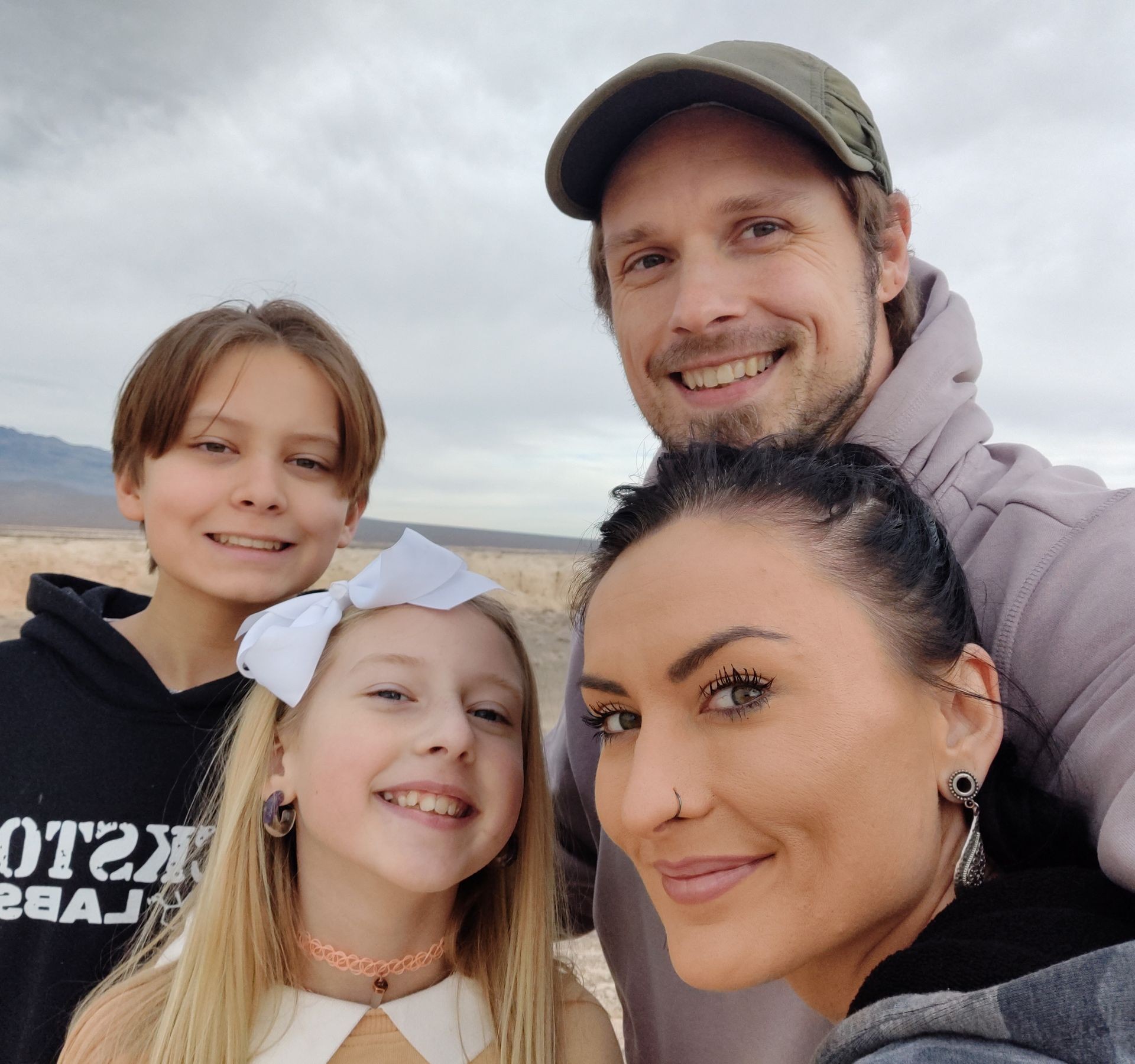 The image features a happy family of four smiling at the camera. The father, wearing a cap, has his arm around the eldest child, a young teenager. The mother, prominently featured in the foreground, is sporting a sleek hairstyle and a nose ring, and has a bright smile. The youngest child, with a large white bow in her hair, is in front of the group. They all appear to be enjoying a day out in nature.